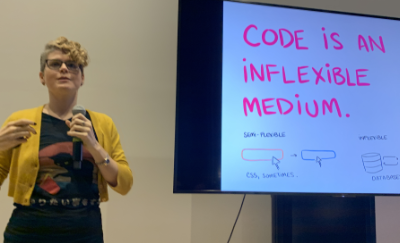 An image of CJ standing in front of a slide that says, 'Code is an Inflexible Medium.'